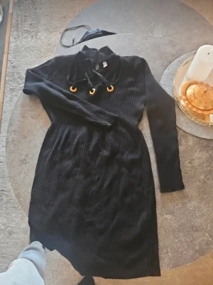 Svart ribbad klänning med snörning - Svart ribbad klänning med lång ärm och snygg snörning med guldfärgade ringar vid halsen. Klänningen har en hög krage och är i ett stretchigt stickat material som sitter snyggt på kroppen.