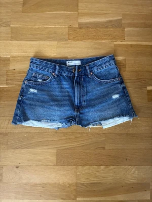 Blå jeanskjol från Bershka strl 36 / XS - Säljer en kort blå jeanskjol från Bershka med rå kant och slitna detaljer. Kjolen har klassisk femficksdesign och bälteshällor. Perfekt för dig som gillar en avslappnad och trendig stil.