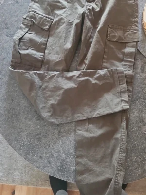Olivgröna cargobyxor med fickor - Säljer ett par olivgröna cargobyxor med flera stora fickor på sidorna och bak. Byxorna har en avslappnad passform och är tillverkade i ett slitstarkt bomullsmaterial. Perfekta för dig som gillar streetwear och praktiska detaljer.