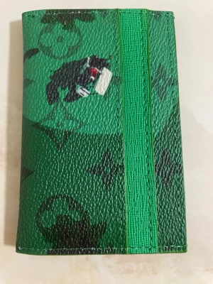 Korthållare - Snygg grön korthållare med klassik monogrammönster och coola svarta detaljer. Tillverkad i tåligt läder med flera fack för kort och sedlar. Perfekt för dig som vill ha något unikt och trendigt i fickan.