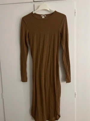 Brun långärmad klänning - En enkel och stilren brun långärmad klänning med rund halsringning. Klänningen har en rak passform men sitter ändå efter ens egna kroppsform och är tillverkad i ett mjukt material som känns skönt mot huden. Perfekt för dig som gillar minimalistisk stil.