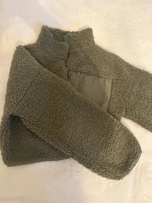 Grön teddytröja med zip - Mysig grön teddytröja med hög krage och zip framtill. Tröjan har en praktisk bröstficka med dragkedja och är perfekt för kyliga dagar. Materialet är fluffigt och stickat för extra komfort.