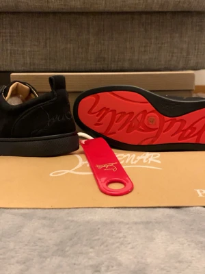 Svarta Louboutin sneakers med röd sula - Stilrena svarta sneakers från Christian Louboutin i mocka med ikonisk röd sula och diskret broderad logga på hälen. Lågt skaft och klassisk siluett som passar till många olika looks. Kommer med originaltillbehör.