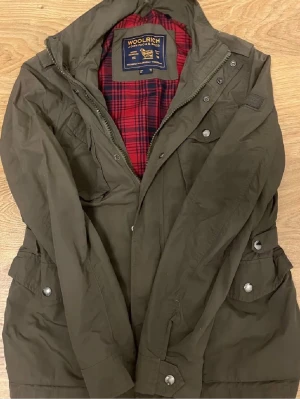  Field jacka Woolrich - Snygg olivgrön parkajacka från Woolrich med fyra stora fickor framtill och dragkedja samt knappar. Jackan har ett rött rutigt innerfoder och hög krage. Perfekt för dig som vill ha en klassisk och funktionell jacka med coola detaljer.