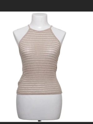 Beige virkad halterneck topp - Trendig beige halterneck topp med virkad look och smala axelband. Toppen har ett genombrutet mönster som ger en luftig känsla och är perfekt för sommaren. Materialet ser ut att vara bomull eller en bomullsblandning.