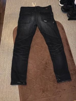 Svarta Hugo Bosss slim fit jeans med slitningar - Säljer ett par svarta Hugo Boss slim fit jeans med snygga slitningar och tvättade detaljer baktill. Jeansen har raka ben och två bakfickor. Perfekta för en avslappnad och trendig look.