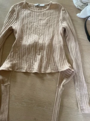 Beige ribbad topp med knytband - Trendig beige långärmad topp med ribbad struktur och breda knytband i midjan som kan knytas bak. Toppen har rund halsringning och är croppad i modellen, perfekt för en snygg och avslappnad look.