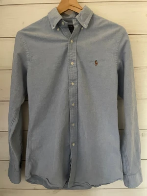 Blå skjorta från Polo Ralph Lauren - Snygg ljusblå skjorta från Polo Ralph Lauren med klassisk button-down krage och broderad logga på bröstet. Materialet är mjukt och känns bekvämt mot huden.