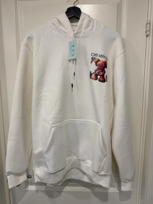 Vit Off-White hoodie med tryck - Säljer en vit hoodie från Off-White med ett färgstarkt tryck av en KAWS-figur på bröstet och Off-White-loggan ovanför. Hoodien har huva, känguruficka och långa ärmar. Perfekt för dig som gillar streetwear och unika prints.