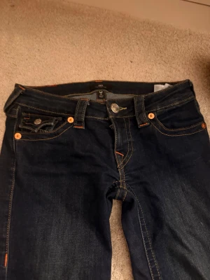 Mörkblå True Religion jeans - Säljer ett par mörkblå jeans från True Religion med kontrastsömmar och snygga detaljer på fickorna. Klassisk femficksmodell med silverfärgade knappar och logopatch bak. Perfekta för dig som gillar streetwear och vill ha ett par jeans med unik design.