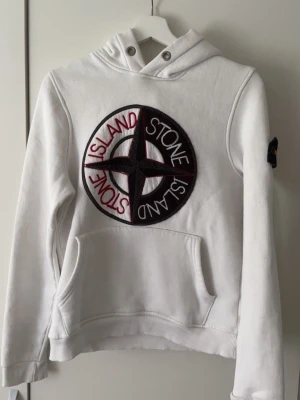 Vit hoodie  - Vit hoodie från Stone Island med stor broderad logga i svart, rött och vitt på bröstet samt klassisk patch på ärmen. Hoodien har huva med dragsko och känguruficka framtill. Tillverkad i mjuk bomull för skön känsla.