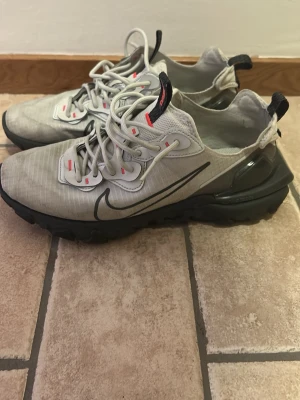 Nike React Element 55 sneakers grå - Nike React Element 55 sneakers i ljusgrå mesh med svarta detaljer och reflexer. Skorna har en chunky svart sula med vågig design, vita snören och små röda accenter. Swoosh-loggan syns tydligt på sidan. Perfekta för dig som gillar sportig och modern stil.