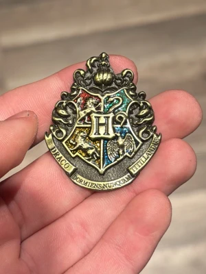 Hogwarts Emblem I Harry Potter - Metallpin med Hogwarts vapensköld från Harry Potter. Skölden har detaljer i rött, grönt, blått och gult. Perfekt accessoar för samlare eller entusiaster. Finns en mindre skada på emblemet som tydligt ses på bilden, den märks inte för andra än dig själv.