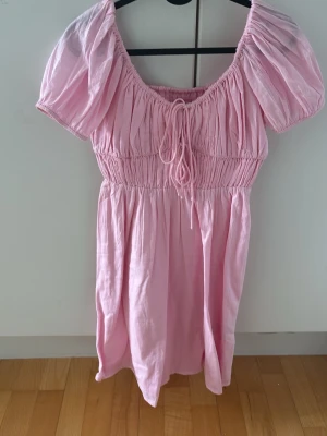 Rosa kort puffärmad klänning - Söt rosa kortklänning med puffärmar och smock i midjan. Klänningen har en rundad urringning med knyt framtill och är tillverkad i ett mjukt bomullsliknande material. Perfekt för en somrig look med voluminösa ärmar och avslappnad passform.