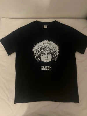 Khabib t shirt - “Smesh” - Cool svart t-shirt med ett stort vitt tryck av ett ansikte med lockigt hår och texten 'SMESH' under. T-shirten har rund hals och korta ärmar. Perfekt för dig som gillar unika och grafiska motiv. Materialet känns som mjuk bomull.