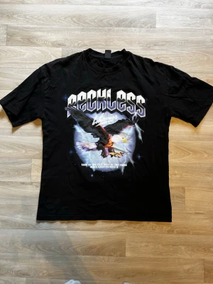 Svart Reckless t-shirt med örntryck - Cool svart t-shirt med stort tryck framtill som visar en örn och texten 'Reckless'. T-shirten har klassisk passform och rund hals. Perfekt för dig som gillar streetwear och grafiska motiv.