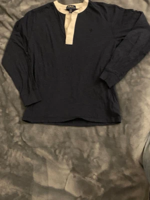 Mörkblå långärmad topp Polo Ralph Lauren - Mörkblå långärmad topp från Polo Ralph Lauren med kontrasterande beige knäppkant och liten broderad logga på bröstet. Tillverkad i mjuk bomull och har en avslappnad passform. Perfekt för en chill och stilren look.