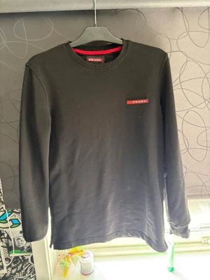 Svart sweatshirt från Prada - Snygg svart sweatshirt från Prada med rund halsringning och diskret röd logotyp på bröstet. Tröjan har långa ärmar och ribbade muddar vid hals, ärmslut och nederkant. Perfekt för en clean och stilren look.