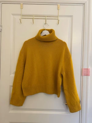 Gul polotröja i stickat material - Säljer en snygg gul stickad polotröja med breda ribbade muddar och hög krage. Tröjan har en boxy passform och långa ärmar, perfekt för att hålla värmen under kyliga dagar. Materialet känns mjukt och mysigt, och färgen är riktigt poppig.