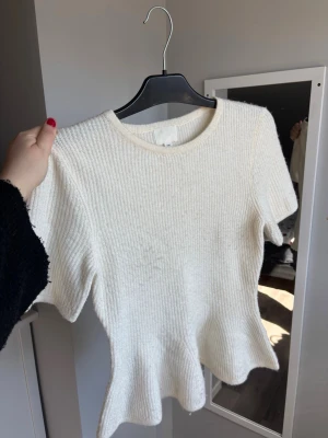 Vit ribbad topp med peplum - Supersöt vit ribbad topp med korta ärmar och peplum nedtill. Mjuk och stretchig känsla, perfekt till jeans eller kjol. Rund halsringning och figurnära passform som framhäver midjan.