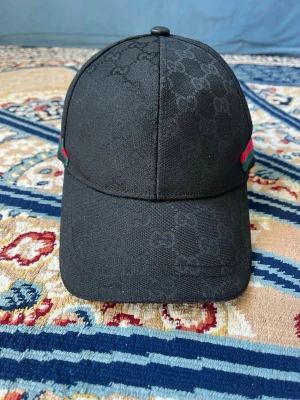 Svart keps från Gucci med GG-mönster - Snygg svart keps från Gucci med det klassiska GG-monogrammet ton-i-ton över hela kepsen. På sidorna finns gröna och röda detaljer som ger en exklusiv känsla. Kepsen har böjd skärm och knapp på toppen. Perfekt accessoar för en trendig look.