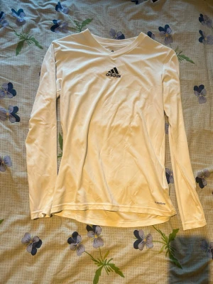 Vit långärmad träningströja Adidas - Vit långärmad träningströja från Adidas med svart logga på bröstet och diskret 'Climacool' tryck vid nederkanten. Tillverkad i ett lätt och ventilerande funktionsmaterial, perfekt för träning och sport.