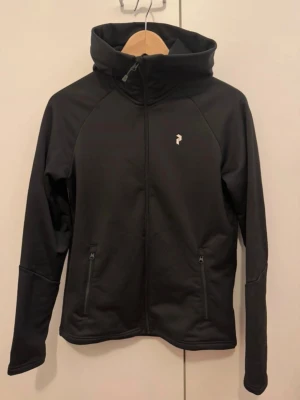 Peak Performance rider hood - Snygg svart kofta från Peak Performance med dragkedja framtill, två fickor med dragkedja och huva. Logotyp på bröstet och stor Peak Performance-text på huvan. Jackan har en sportig passform och är tillverkad i ett lätt, vindtätt material.