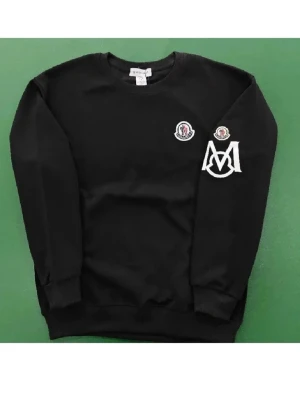 Moncler sweatshirt - Säljer en svart sweatshirt från Moncler med rund halsringning och ribbade muddar. Tröjan har vita och röda Moncler-loggor på bröstet och ärmen samt en stor vit M-brodyr på ena ärmen. Perfekt för dig som gillar streetwear och snygga detaljer.