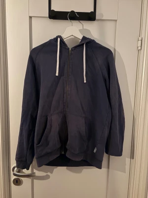 Mörkblå zip hoodie - En enkel och stilren mörkblå hoodie med dragkedja framtill och vita snören i huvan. Hoodien har två stora fickor på magen och ribbade muddar vid ärmslut och nederkant. Perfekt för en avslappnad look.