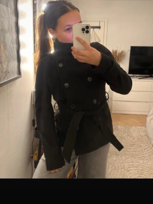 Svart trenchcoat med knytskärp - Stilren svart trenchcoat med dubbelknäppning och brett knytskärp i midjan. Jackan har hög krage och raka ärmar, vilket ger en klassisk och clean look. Passar perfekt till jeans eller kjol för en trendig outfit.