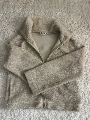 Beige teddyjacka med dragkedja - Mysig beige teddyjacka med hög krage och dragkedja framtill. Jackan har en fluffig, mjuk yta och raka ärmar. Perfekt för att hålla värmen under kyliga dagar. Enkel och stilren design utan synligt märke.