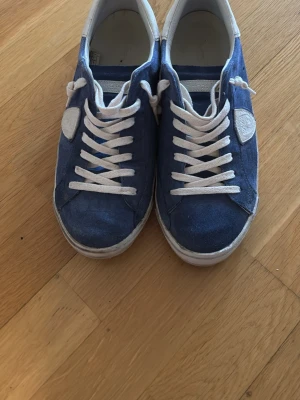 Blåa sneakers från Philippe Model - Snygga blåa sneakers från Philippe Model med vita detaljer, vita skosnören och vit sula. Skorna har en låg profil och är tillverkade i mocka med läderdetaljer på häl och sidor. Perfekta för en avslappnad och trendig stil. Kanske använt 10 gånger! 