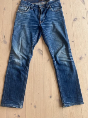Blå jeans från Nudie Jeans - Säljer ett par klassiska blå jeans från Nudie Jeans med snygg slitning och raka ben. Jeansen har fem fickor, orangea sömmar och patch bak i midjan. Tillverkade i denim av bomull. Perfekta för en avslappnad och trendig look.
