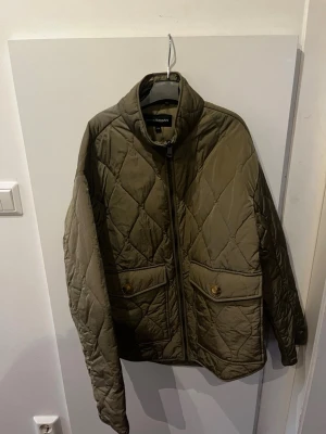 Olivgrön quiltad pufferjacka KappAhl - Snygg olivgrön pufferjacka från KappAhl i storlek XS. Jackan har quiltat mönster, hög krage och dragkedja framtill. Två stora fickor med knappstängning ger extra stil och funktion. Perfekt för kyliga dagar och enkel att matcha med olika outfits. Köpt begagnad men aldrig använd av mig. Skulle säga att den är ljusare i verkligheten 