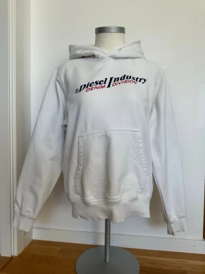 Vit hoodie från Diesel Industry - Snygg vit hoodie från Diesel Industry Denim Division med tryck i marinblått och rött på bröstet. Klassisk känguruficka framtill och huva. Tillverkad i mjukt bomullsmaterial som känns skönt mot huden. Perfekt för en avslappnad streetwear-look. Hoodien har subtila fläckar på armen, framtill och luvan men det är inget som förstör helhetsintrycket.