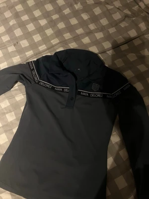 Svart långärmad polotopp Maya Delorez - Snygg svart långärmad polotopp från Maya Delorez med logga och textdetaljer längs bröstet. Toppen har krage och knäppning framtill, perfekt för en sportig och stilren look.