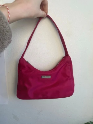 Rosa axelremsväska från Don Donna - Trendig rosa handväska från Don Donna med axelrem och silverfärgad logga framtill. Väskan har en enkel, stilren design och är tillverkad i ett glansigt tygmaterial. Perfekt för att lyfta din outfit med en färgklick.