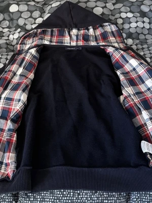 Leon Marcel marinblå hoodie med rutigt foder - Snygg marinblå hoodie från Marcel med dragkedja och huva. Insidan av huvan och öppningen har ett rutigt foder i rött, vitt och blått. Broderad logga på bröstet och ribbade muddar vid ärmar och nederkant. Snören från huvan är uttagna. Storlek M men sitter som S