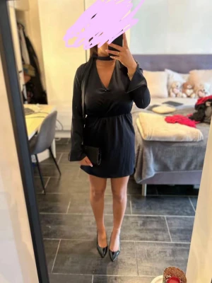 Mörkblå omlottklänning med v-ringning - Stilren svart kortklänning med omlottdesign och djup v-ringning. Klänningen har långa ärmar och markerad midja, vilket ger en smickrande siluett. Perfekt för dig som gillar en elegant och enkel look.