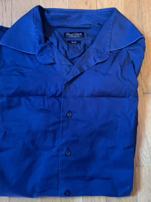 Marinblå skjorta från David Saddler - Snygg marinblå skjorta från David Saddler i slim fit-modell. Skjortan har klassisk krage, knappar framtill och är tillverkad i ett mjukt bomullstyg. Perfekt för dig som gillar stilrena och enkla plagg.