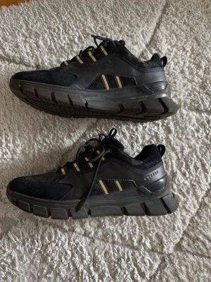 Svarta sneakers från Court - Tuffa svarta sneakers från Court med chunky sula och detaljer i mocka och textil. Skorna har svarta och beige ränder på sidan samt snörning. Perfekta för dig som gillar en sportig och modern stil.