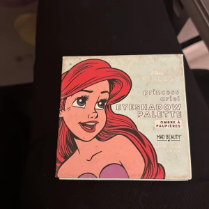 Disney Princess Ariel ögonskuggspalett - Ögonskuggspalett inspirerad av Disney Princess Ariel från Mad Beauty. Paletten innehåller nio färgstarka nyanser i lila, turkos, grön, guld, rosa, orange och röd, både matta och skimrande. Perfekt för kreativa och lekfulla sminkningar.
