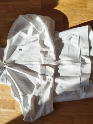 Vit hoodie från Polo Ralph Lauren - Säljer en vit hoodie från Polo Ralph Lauren med dragkedja framtill och klassisk broderad logga på bröstet. Hoodien har fickor på sidorna och en skön huva. Tillverkad i mjukt bomullsmaterial som är perfekt för chill dagar.