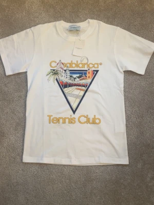 Vit Casablanca Tennis Club t-shirt - Snygg vit t-shirt från Casablanca med färgglatt tryck framtill som visar en tennisbana och byggnader. Texten 'Casablanca Tennis Club' i gult ger en retrovibe. Klassisk passform och rund hals, perfekt för en avslappnad look. Lite stor i storlek så passar även M.