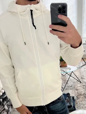 Vit hoodie CP company - Snygg vit hoodie från CP company med dragkedja framtill och två fickor med dragkedja. Klassisk huva med dragsko och diskret logga på dragkedjan. Perfekt till jeans eller joggers för en clean och stilren look.