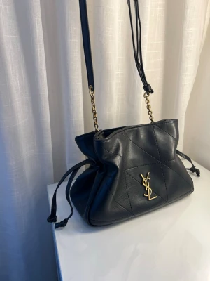 Svart YSL handväska i läder - Snygg svart handväska från Yves Saint Laurent i mjukt läder med quiltade detaljer. Väskan har guldfärgad YSL-logga framtill, justerbar axelrem i läder och kedja samt knytband på sidorna. Kom gärna privat för fler bilder eller frågor.