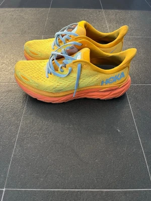 Hoka Clifton 8 gula sneakers - Färgglada Hoka Clifton 8 sneakers i gult mesh med orange sula och ljusblå skosnören. Lätta och ventilerande med en chunky sula för extra komfort. Perfekta för löpning, vandring och träning. Använda få gånger. Nypris 1799. Pris går att diskutera. Passar storlek 40 1/2 - 41. 