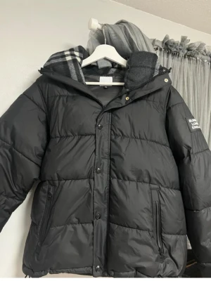 Svart pufferjacka Burberry London - Snygg svart pufferjacka från Burberry London med huva och dragkedja samt knappar framtill. Jackan har två fickor med dragkedja och ett diskret Burberry-tryck på ärmen. Fodret har klassiskt rutmönster i grått och vitt. Perfekt för kalla vinterdagar.