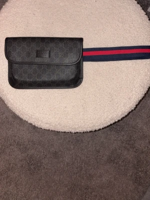 GG svart bälte med väska - Snyggt svart bälte från GG med tillhörande väska i skinn och klassiskt GG-mönster. Bältet har marinblå och röda ränder som ger en ikonisk look. Perfekt accessoar för att lyfta din outfit och ha det viktigaste nära till hands.