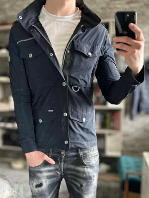 Snoor Jacka - Snygg mörkblå vindjacka med flera framfickor, silverfärgade knappar och dragkedja. Jackan har lång ärm, hög krage och detaljer som D-ring och bröstficka med dragkedja. Perfekt för dig som gillar en sportig och stilren look.